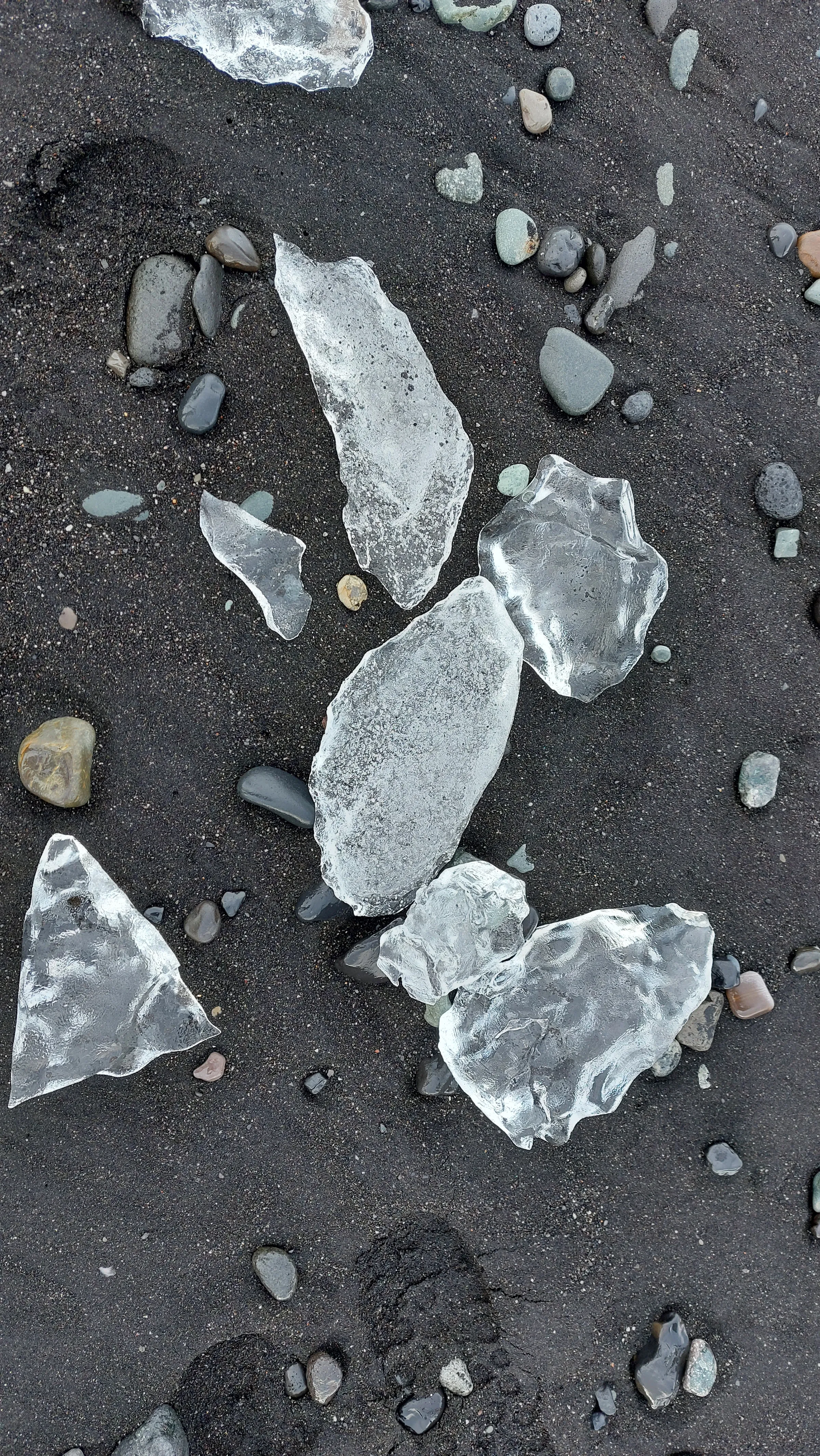 Kristalheldere ijsbrokken tussen donkere kiezels op zwart zand.